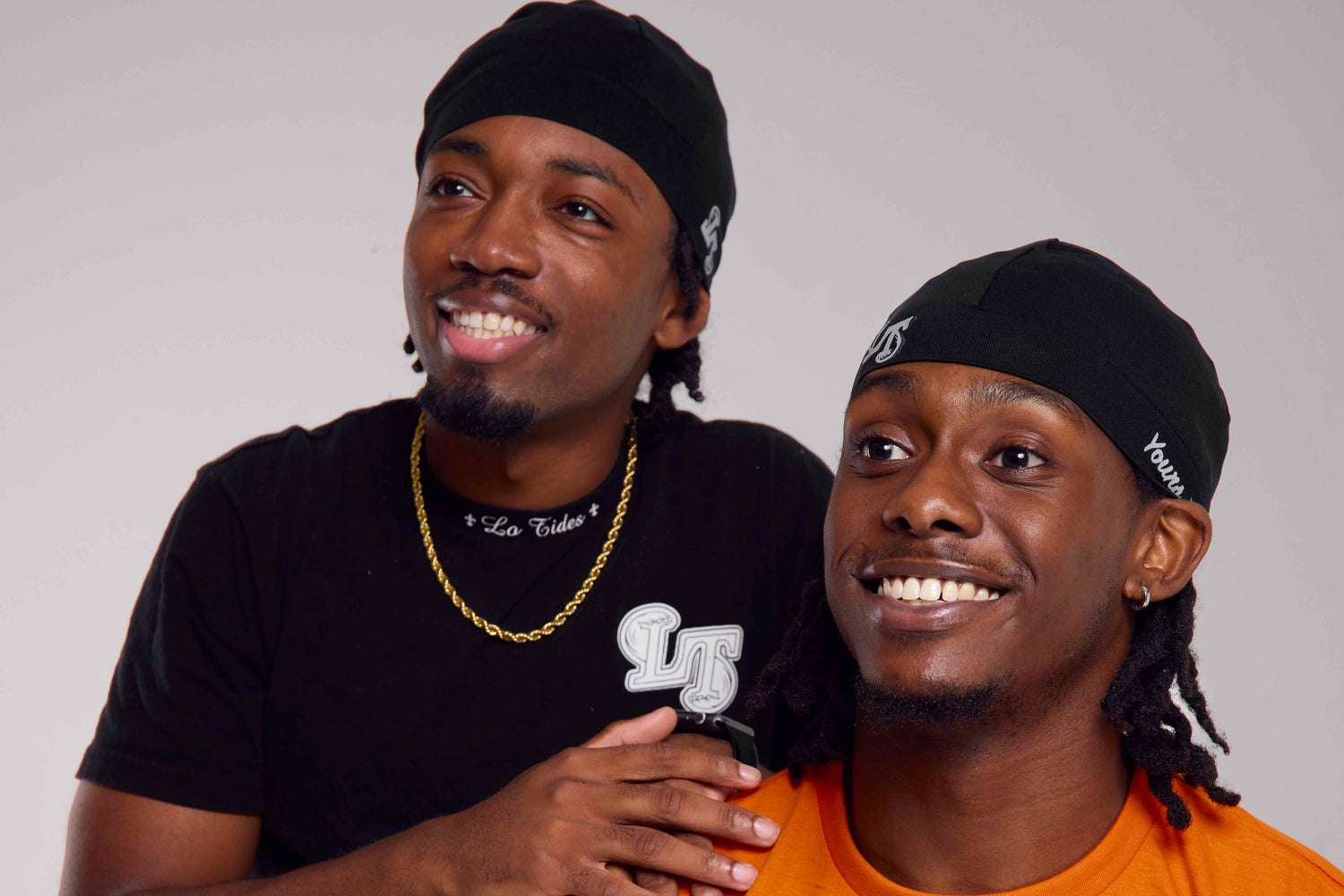 Two men wearing black caps with logos, one in a black shirt and the other in an orange shirt, on a plain background.
