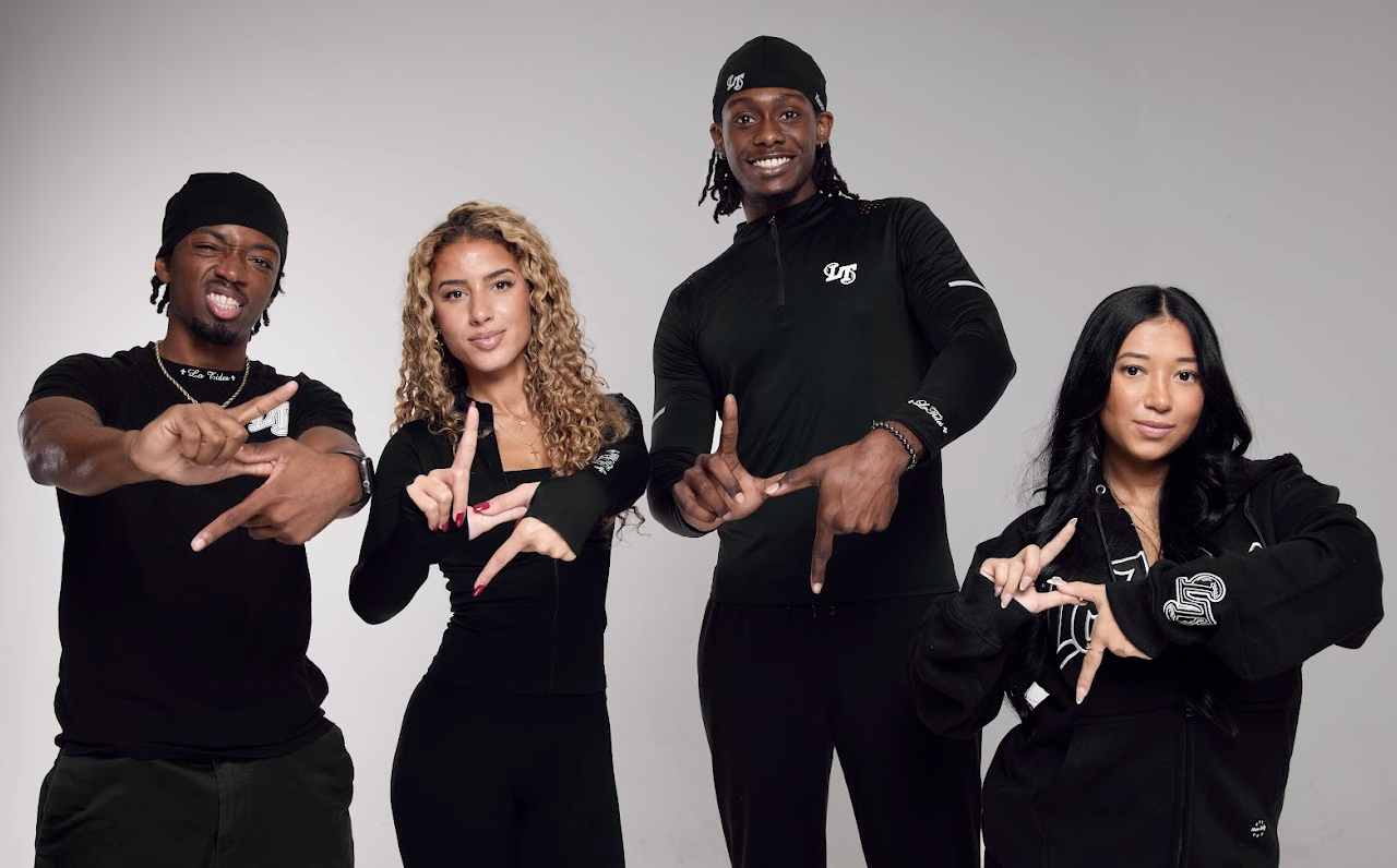 Four people wearing black outfits with logos, posing against a gray background